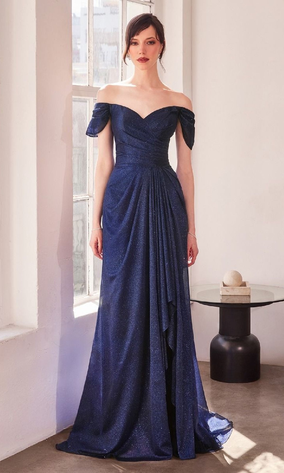 A woman stands indoors next to a small round table, wearing the Off-the-Shoulder Glitter Prom Dress: Amoris T604, featuring draped detailing and a floor-length skirt.